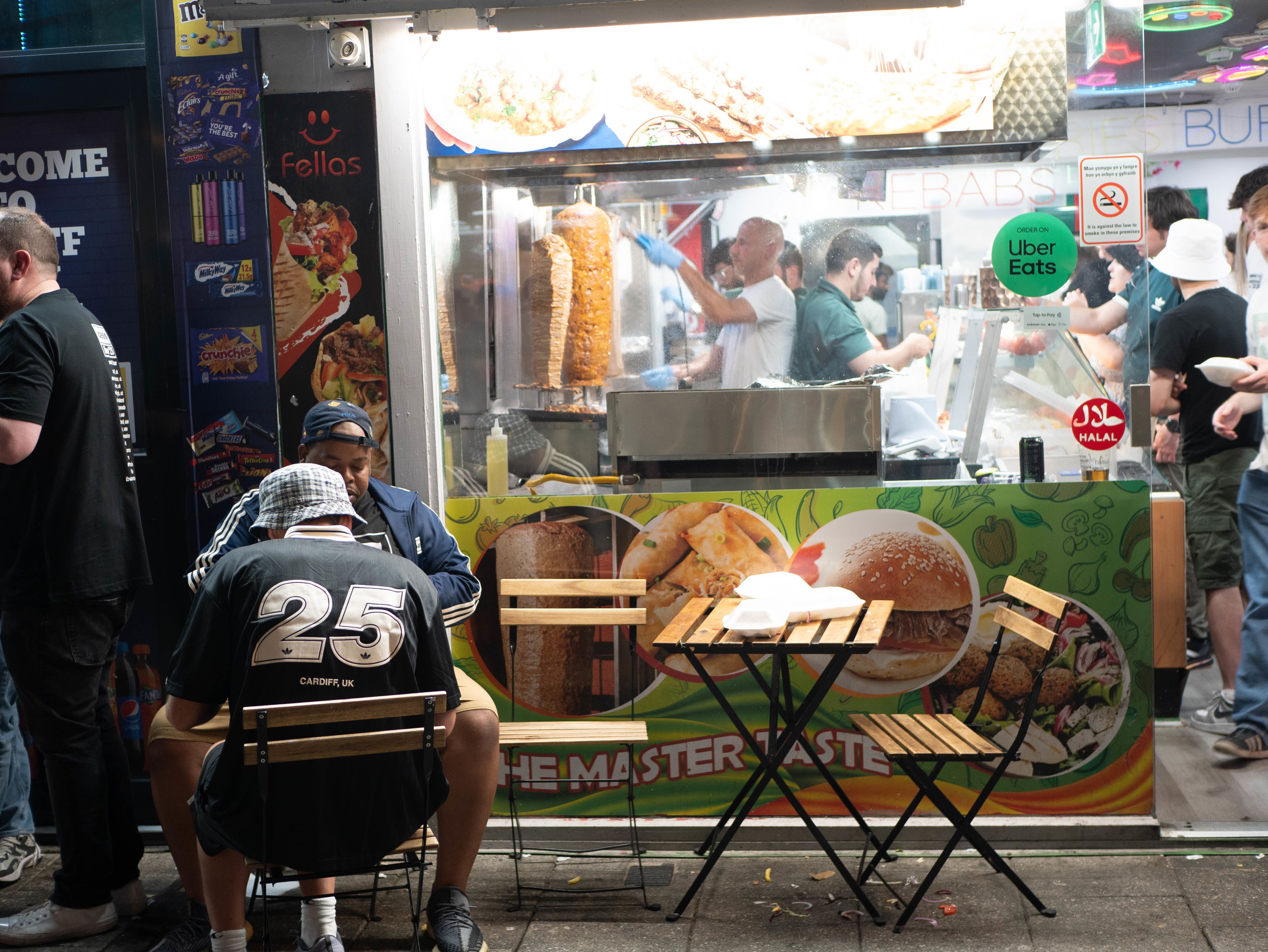 Two men in Oasis merchandise eating outside of crowded kebab shop on Caroline Street